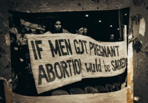 A black and white photograph, taped to a pole, depicting a banner reading "IF MEN GOT PREGNANT ABORTION WOULD BE SACRED"
