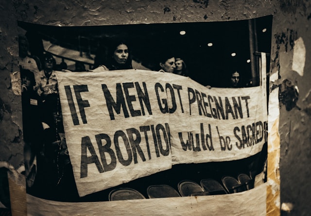 A black and white photograph, taped to a pole, depicting a banner reading "IF MEN GOT PREGNANT ABORTION WOULD BE SACRED"