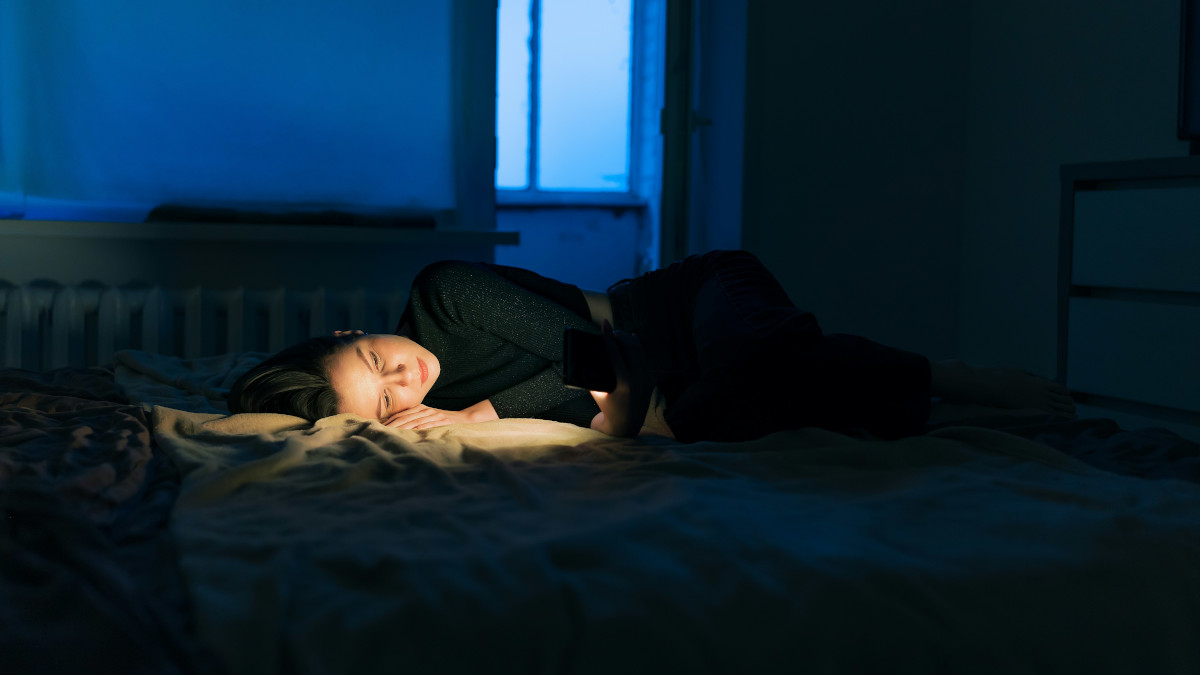 Woman staring at her phone screen on her bed with the lights turned off